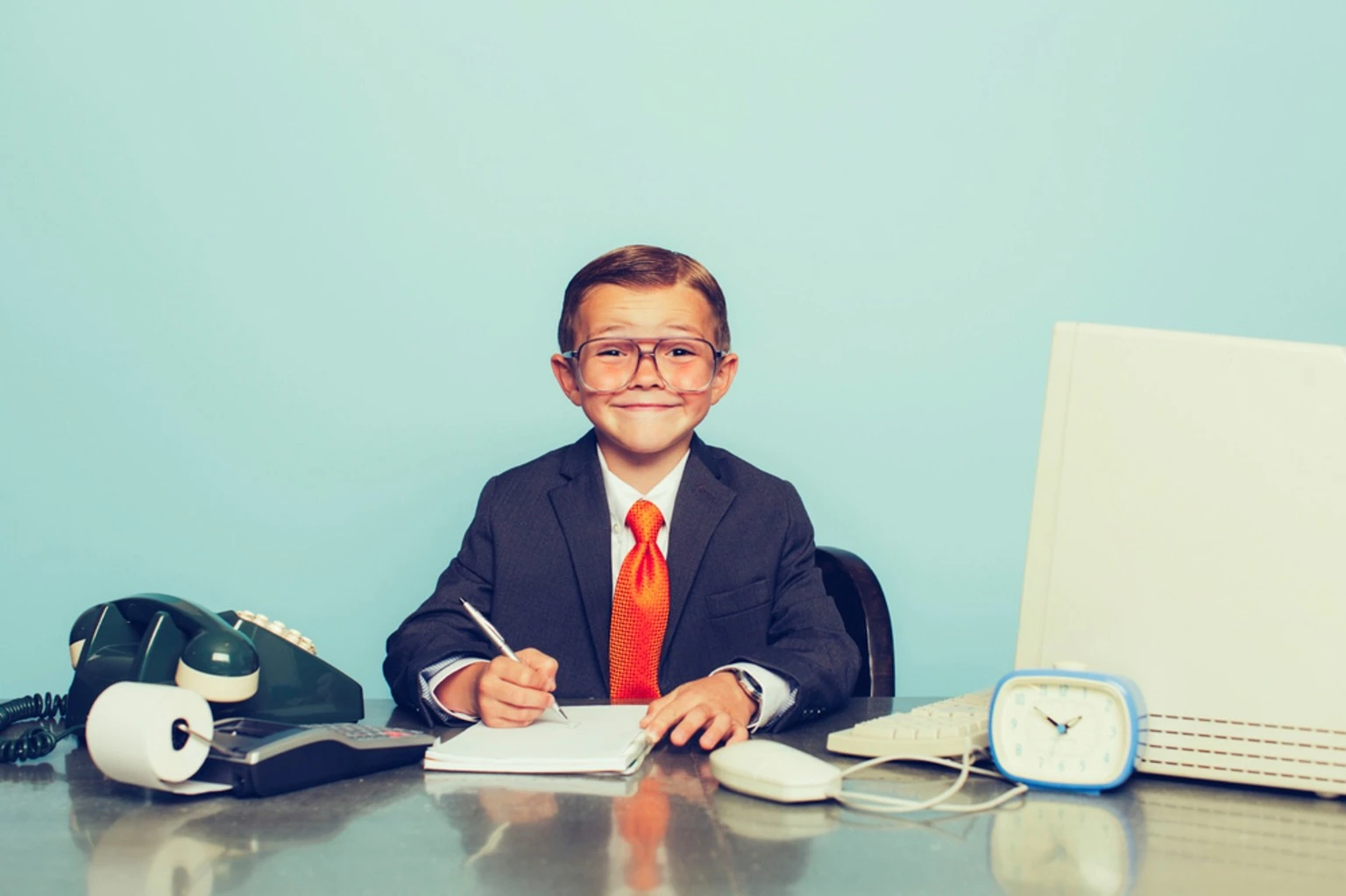 Un niño con traje y corbata trabaja entre una calculadora, un teléfono y una computadora vintage
